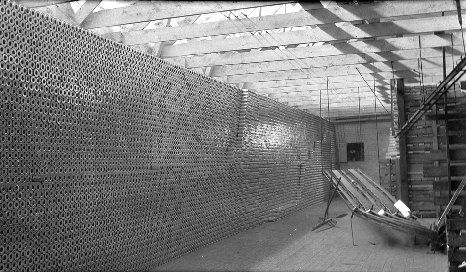 Black and white photo of a huge wall of cans at the Empson cannery. Cans are empty, with a hole in the top, and stacked on their sides across a large room. On the right side of the photo is a conveyor system and additional cans in a wooden rack. The room's ceiling is open to wooden beams and a large skylight, and sunlight fills the room.