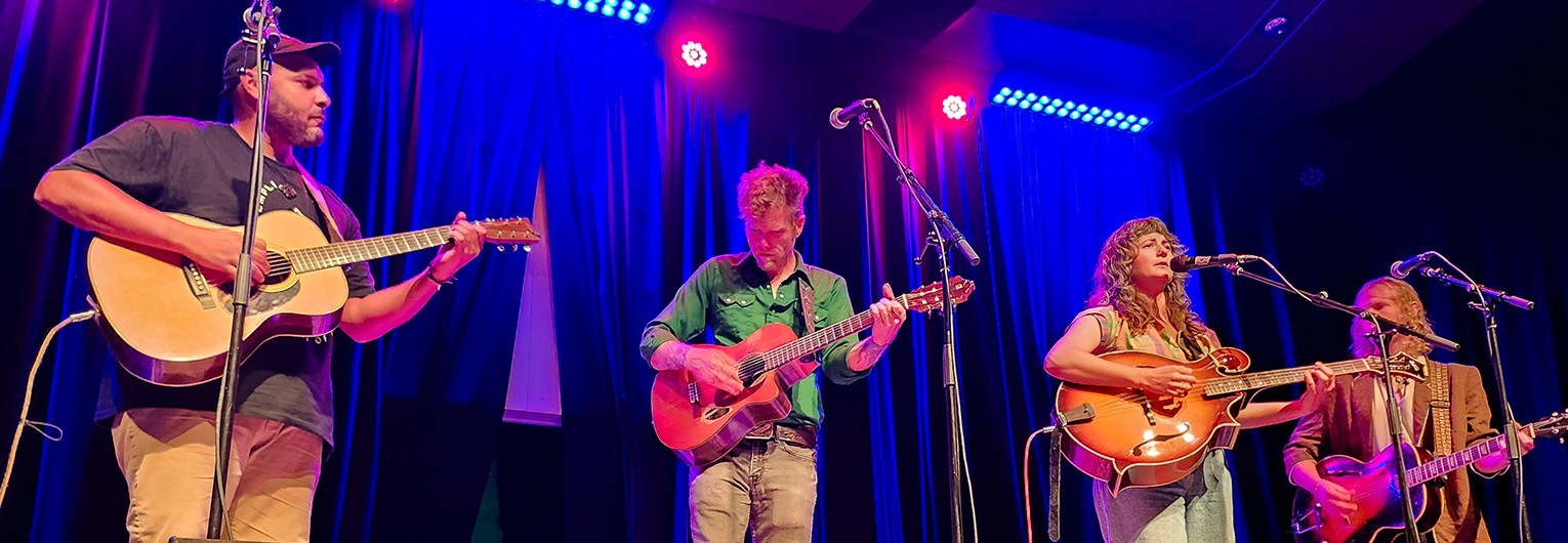 Four musicians on a stage playing instruments against a blue background