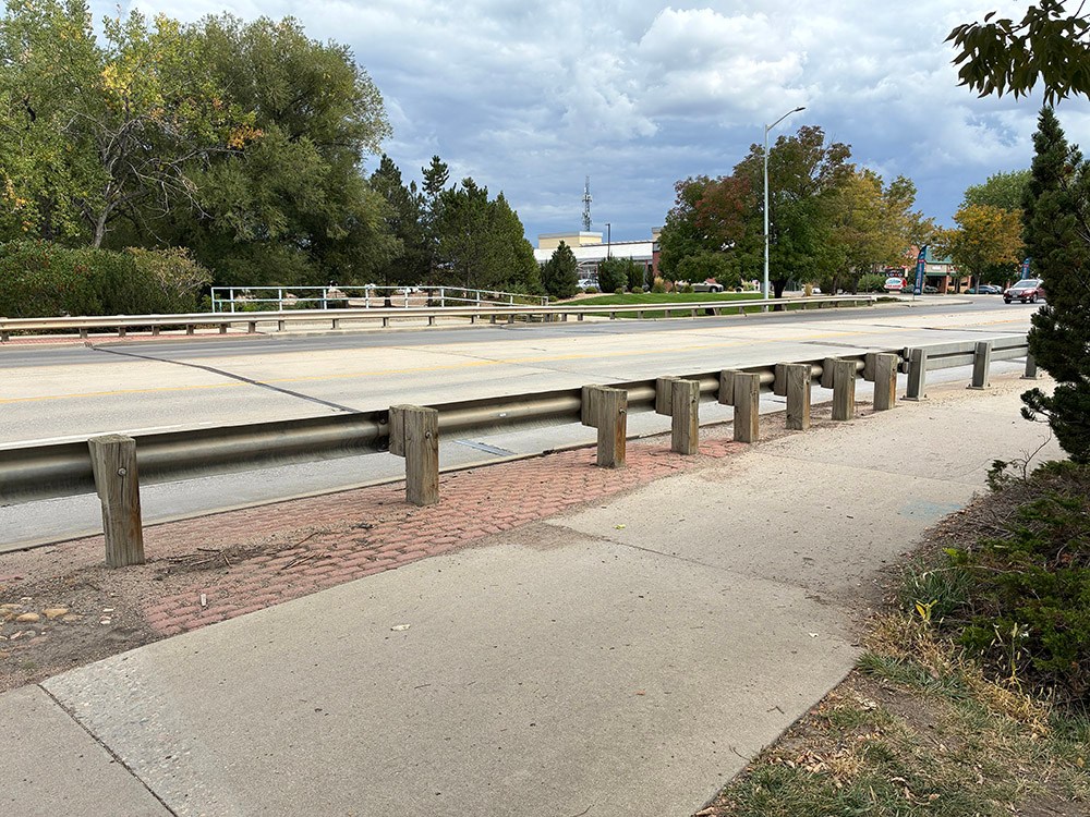 The street side view of a bridge with metal barriers on each side of the road. There are large leafy green trees in the background under a cloudy blue sky.