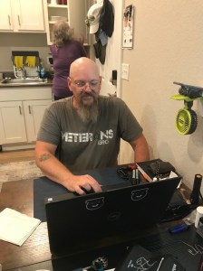 A bearded man with a veterans T-shirt works at a computer on a table. In the background, a woman is opening a cupboard in the kitchen.