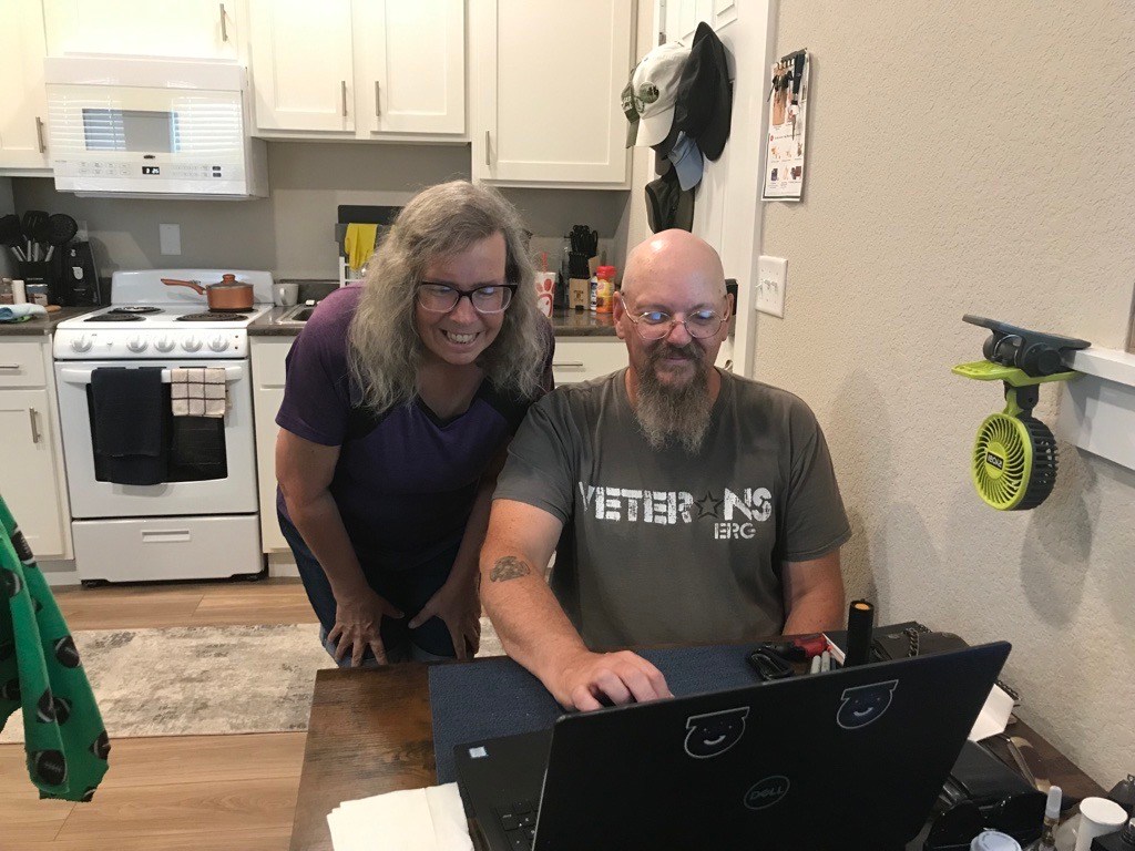 A bearded man and long-haired woman smile as the man works on a computer; the kitchen is visible behind them.