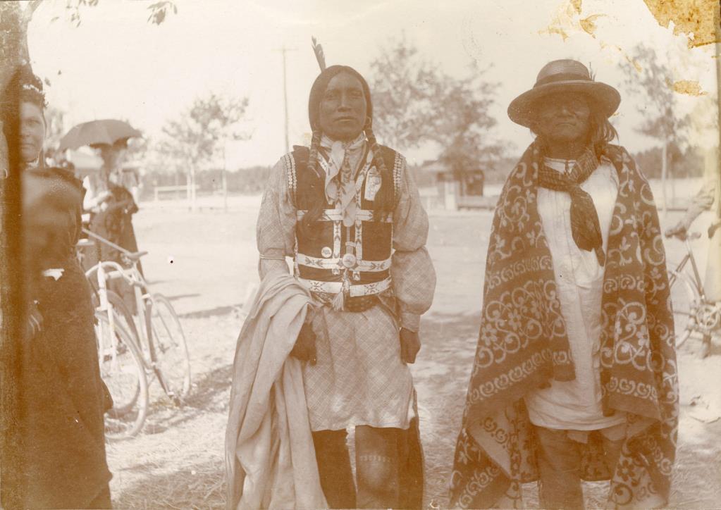 Print of Ute Indians taken at the "Riding Park" (Roosevelt Park). There are two men in the center, dressed in native clothing. The man on the left wears his hair in pigtails, and there is a feather in the back of his hair. The man on the right appears to be much older. He wears a straw hat and a blanket around his shoulders. On the far left is a woman who appears to be Indian. She wears a long, dark-colored dress. Behind her is a man holding an umbrella standing behind a bicycle. Another man on a bicycle is partially visible on the far right. There are trees and a small building in the background.