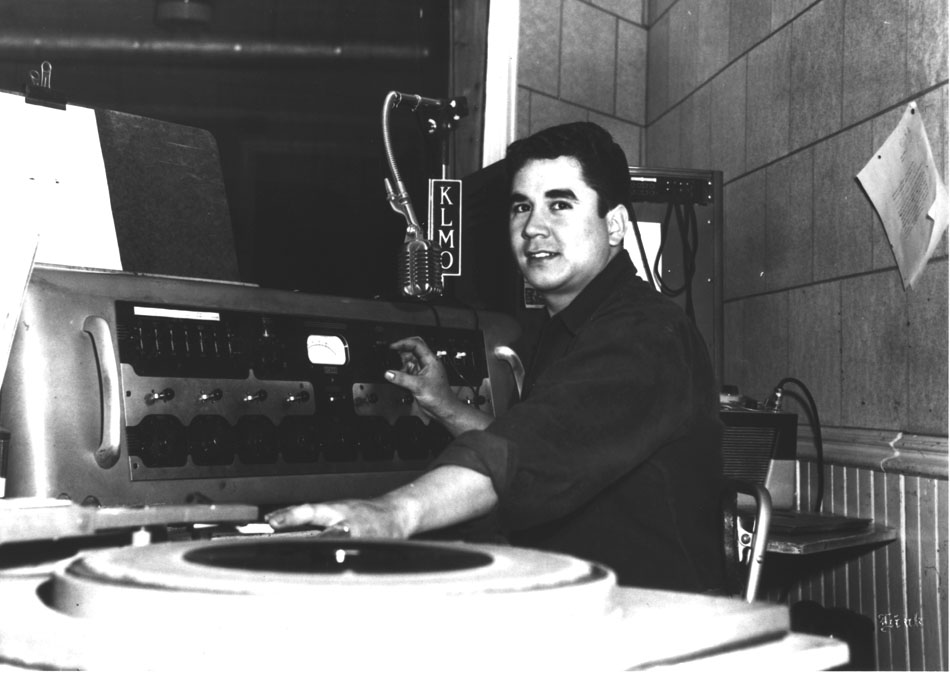 Black and white copy print of Ben Rodriguez at KLMO radio station. Ben is at the controls with a microphone in front of him. He is wearing a dark shirt with sleeves rolled-up.