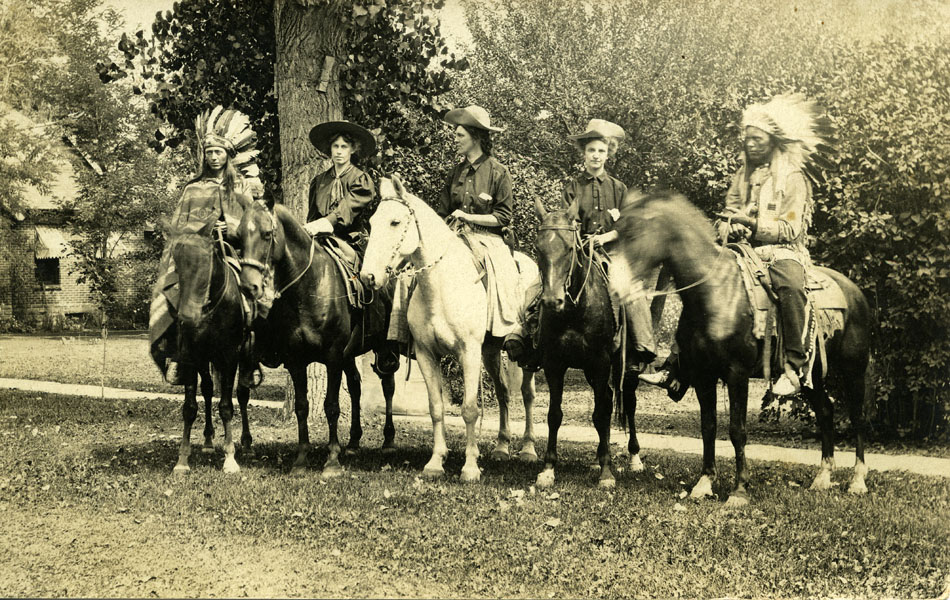 A sepia tone photograph of three women and two native American Indians on horses. Left to right is a male native American Indian with feather headdress wearing a blanket on a dark horse; next is a woman with a large brim dark hat and dark blouse with neck scarf on a dark horse; third is Ellen Wanda McKeirnan McDonald (aka MacDonald) wearing a large upturned brim light color hat, dark shirt and riding chaps; fourth is a woman wearing a light color brim hat, dark blouse with a dark tie on a dark horse; and last on the right is a native American Indian wearing a feather headdress, long sleeve fringed shirt on a dark horse.