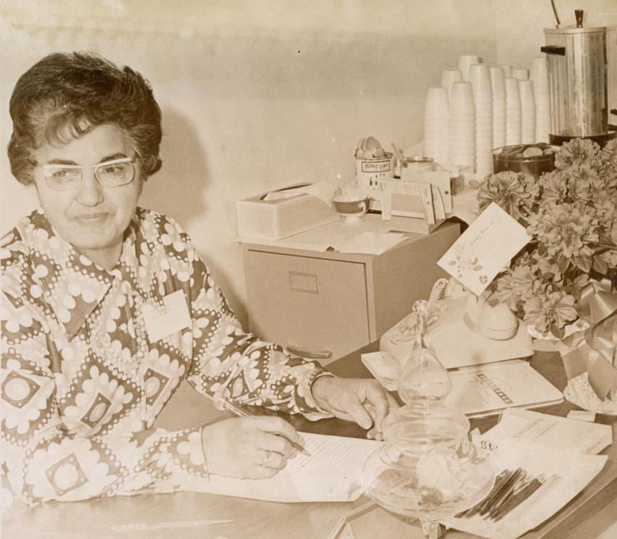 Black and white photo of Eloyeda Cardenas seated at an office desk, with a telephone, a filing cabinet, a bouquet of flowers, a coffee pot, and stacks of coffee cups visible. Caption reads: "Senior citizens are benefitting through the generosity of the First National Bank which is providing them, rent free, a center on Fourth Avenue west of the bank. Here, Eloyada [sic] Cardenas, counselor, is on duty at the front desk to welcome them. Under the OEO, the center provides room for activities, social gatherings, and a place downtown to stopover. Published in the Times-Call on March 27, 1971.