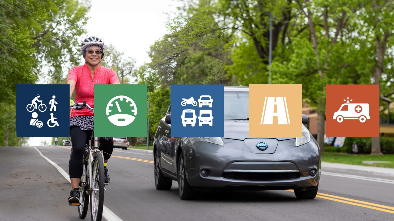 A smiling cyclist in a bright pink jersey, helmet, and yellow gloves rides along a tree-lined street, with a gray electric car approaching behind her. Five colored square icons span the image, representing the Safe System elements: safer people (cyclist, pedestrian, driver, and wheelchair user) in navy, safer speeds (speedometer) in green, safer vehicles (motorcycle, car, bus, truck) in blue, safer roads (road) in tan, and post-crash care (ambulance) in orange. In the lower right is the Vision Zero Longmont logo, "Zero Traffic Deaths."