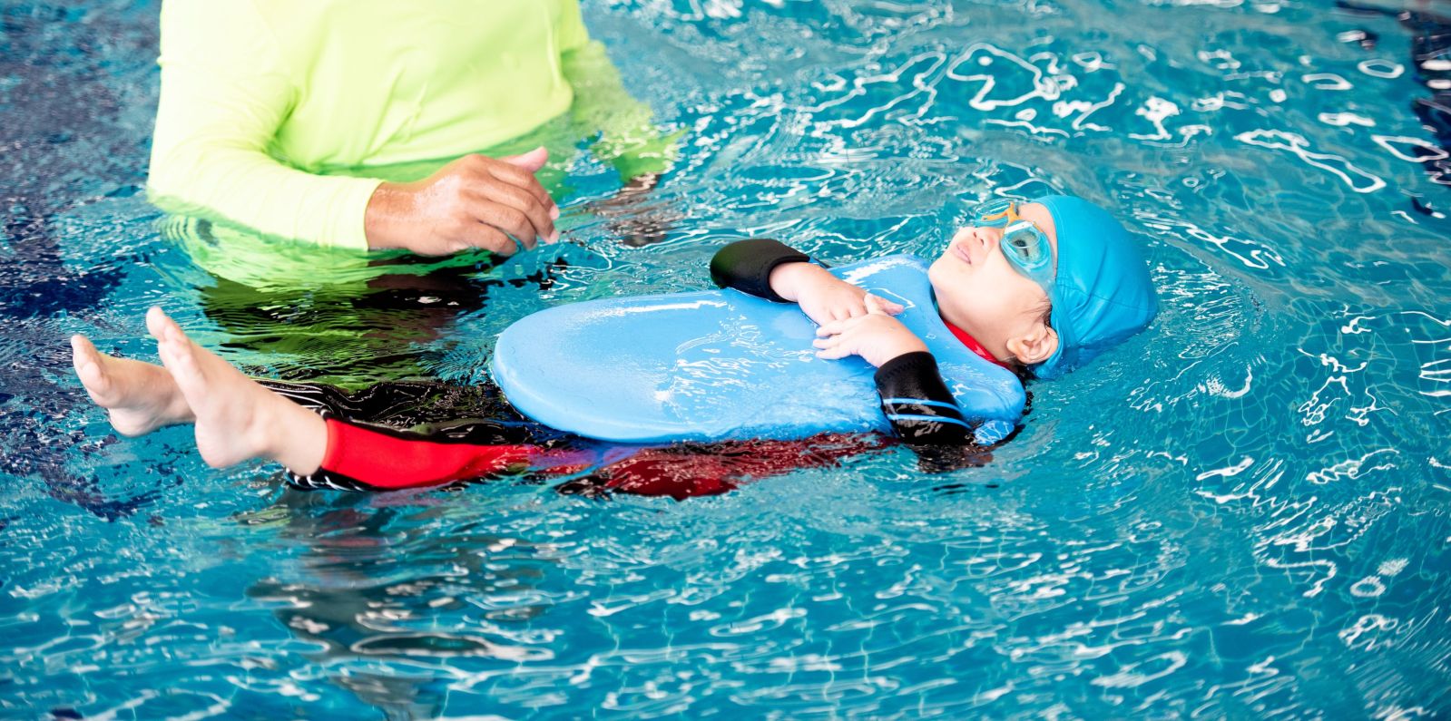 small child swims on their back holding a kick board to help float. An adult is in the water next to him.